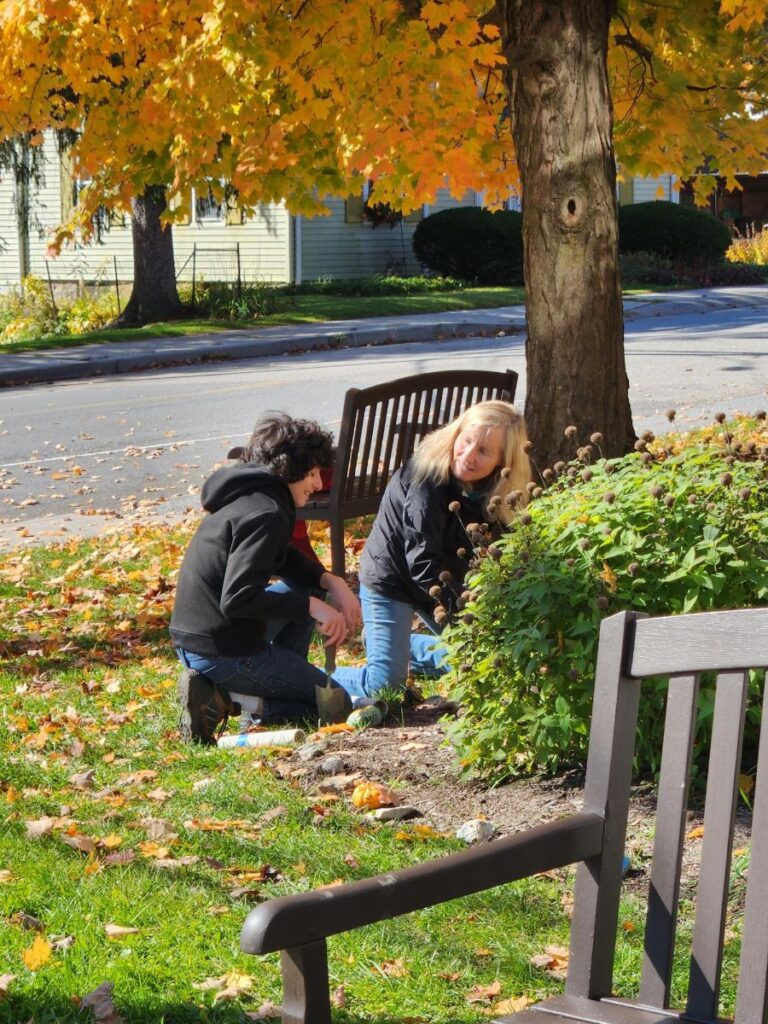 Photo of 2 people kneeling on the ground working in front of a flowering plant