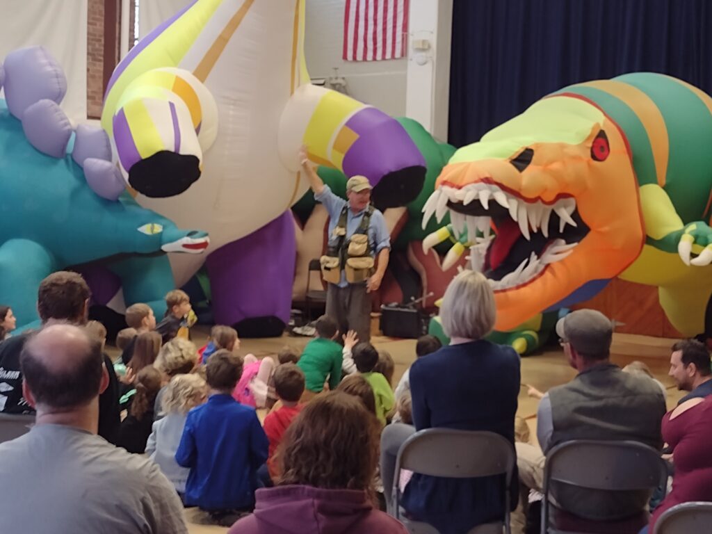 Photo of back of audience, mainly children, looking at a man surrounded by colorful giant blow-up dinosaurs.