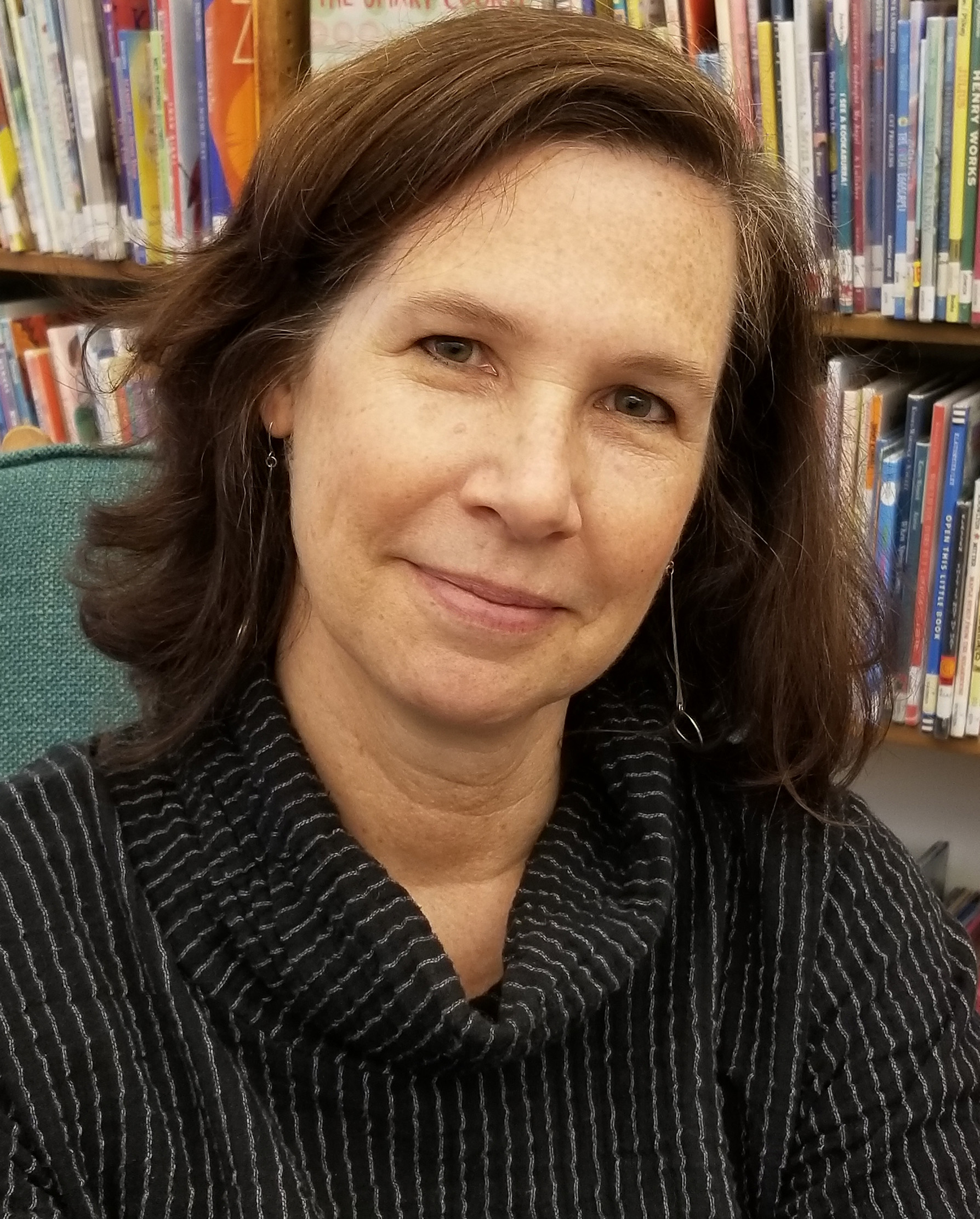 A photograph of Martha Bradford - she is a woman with shoulder-length brunette hair and is sitting in front of a shelf of books.